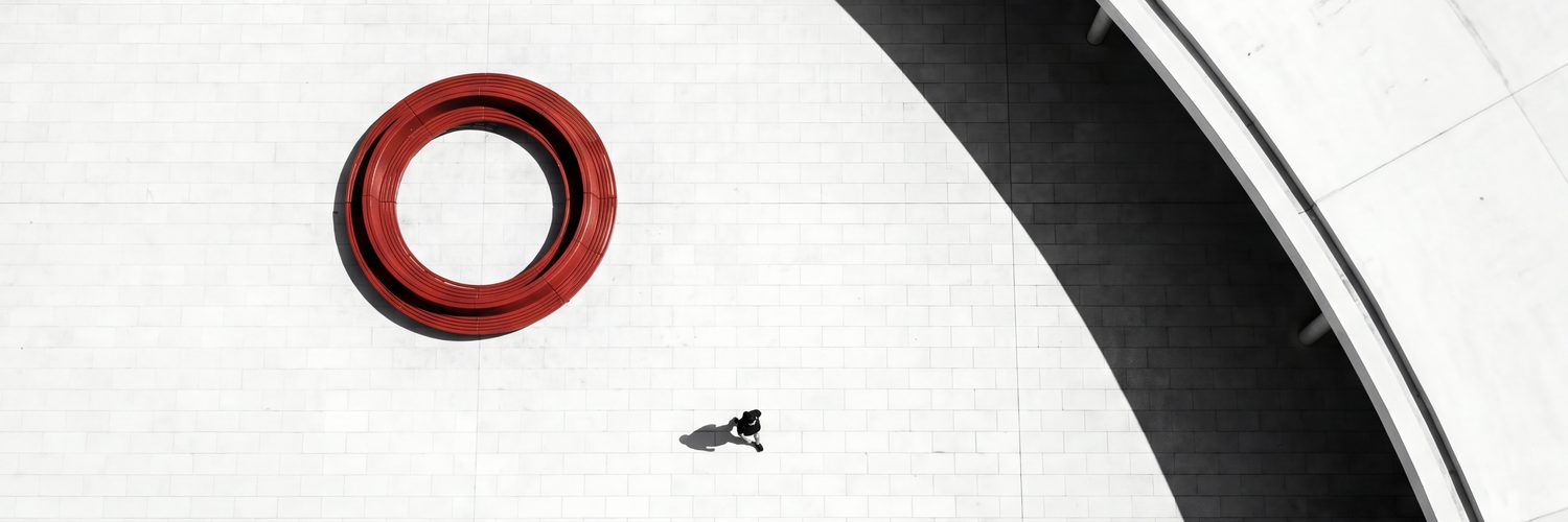 A red circular bench on a pale plaza beside a sweeping curved shadow, one person walking past